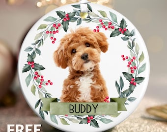 May include: Round ceramic ornament featuring a brown and white dog, surrounded by a wreath of green leaves and red berries. A green banner at the bottom reads "BUDDY". The ornament has a white ribbon for hanging.