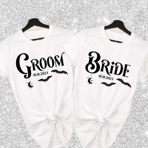 May include: Two white t-shirts with black text. One shirt says "Groom" with the date "01.01.2024" and a crescent moon and bats. The other shirt says "Bride" with the date "01.01.2024" and a crescent moon and bats.