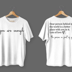 May include: Two white t-shirts with black text. The front shirt says "you are enough". The back shirt says "Dear person behind me, the world is a better place with you in it. Lots of love, -the person in front of you".