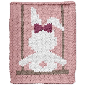 May include: A pink knitted blanket featuring a white bunny design with a burgundy bow tie, sitting on a swing. The swing's supports are brown. The blanket has a textured appearance and a rectangular shape.