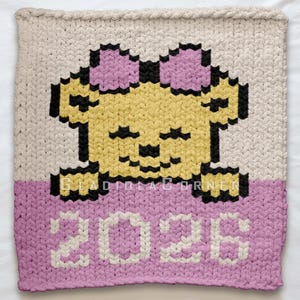 May include: A knitted blanket featuring a pixelated bear design. The bear is yellow with a pink bow and black details. The number "2026" is displayed in white. The blanket has a cream upper section and a pink lower section.