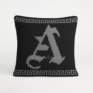 Puede incluir: Un cojín cuadrado de punto negro con una letra A gris grande y centrada en fuente gótica. Los bordes superior e inferior están decorados con un patrón de greca gris horizontal. El texto GLADIOLA CORNER está impreso en letras pequeñas.