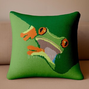 May include: A square knit throw pillow in shades of green featuring a graphic of a red-eyed tree frog with bright orange feet and a light green body. The design includes a diagonal color block effect and the text GLADFUL CORNER in white.
