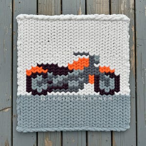 May include: A handmade, square, knitted blanket with a tractor design. The blanket features a white top, a gray bottom, and a tractor in shades of gray, orange, and purple. The text "GLADIOLA CORNER" is visible.