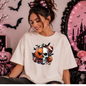 May include: A cream-coloured short-sleeve t-shirt featuring a central graphic design split down the middle. The left side shows an orange jack-o'-lantern, while the right side displays a white human skull, surrounded by footballs and bats.