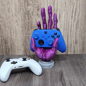 May include: A purple hand-shaped controller holder with a blue controller. A white controller rests on the surface. The holder has a detailed design and a round base. The background is a wooden surface.