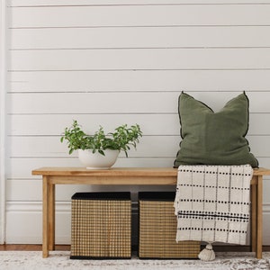 May include: A wooden bench with a green cushion and a white and black throw blanket. Two woven storage baskets are underneath the bench. A potted plant is on the left side of the bench.