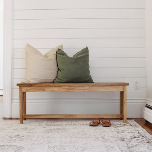 Peut inclure: Un banc en bois avec une finition naturelle, doté de deux coussins, l'un beige et l'autre vert, avec une bordure noire. Le banc est placé devant un mur blanc en lambris et un tapis blanc.