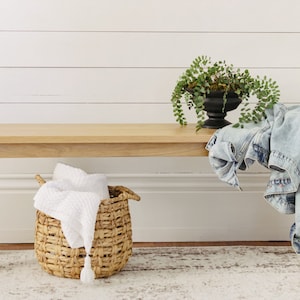 May include: A light wood bench with a white woven basket and a white blanket inside. A denim jacket is draped over the bench. A potted plant with green leaves is on the bench. The bench is in front of a white wall with horizontal wood planks.