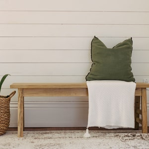 Peut inclure: Un banc en bois avec une couverture blanche et un coussin vert. Le banc est devant un mur blanc en lambris. Une plante en pot est dans un panier tissé à gauche du banc.