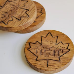 May include: Round wooden coasters with a maple leaf design. The coasters are made of light brown wood. Each coaster features a laser-etched landscape within the maple leaf shape, including trees, mountains, and a bear.