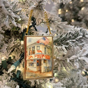 May include: A rectangular Christmas ornament with a watercolor painting of a restaurant with an American flag. The restaurant's sign reads "Lobster Pot Steaks". The ornament is framed in gold and hangs from a gold chain.