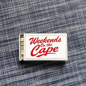 May include: A white matchbox with the words "Weekends On the Cape" in red script. The matchbox is open, revealing the match heads. The background is a blue and white textured fabric.