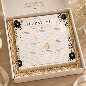 Puede incluir: Una caja de regalo blanca que contiene un bloc de notas Sunday Reset con detalles florales en azul marino y dorado. El planificador incluye secciones para gratitud, bendiciones, lecciones, oración, escrituras y planificación semanal.