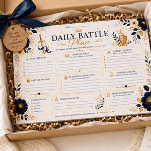 Può includere: Un blocco note Daily Battle Plan in una scatola regalo con un nastro blu navy e un'etichetta rotonda in legno con la scritta You are STRONG and COURAGEOUS. Il blocco bianco presenta decorazioni floreali in oro e blu navy.