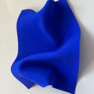 May include: An abstract sculpture in a rich, royal blue. The artwork resembles a draped fabric, with fluid lines and gentle curves. The piece is set against a white backdrop, providing a strong visual contrast.