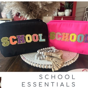 May include: Two black and pink zippered pouches with the word "SCHOOL" in colorful letters. The pouches are on a white beaded surface with a tassel.
