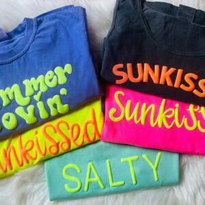 May include: A stack of five t-shirts with different summer-themed slogans. The top shirt is blue with the text "Summer Lovin'" in neon green. The next shirt is yellow with the text "Sunkissed" in orange. The third shirt is pink with the text "Sunkissed" in neon green. The fourth shirt is black with the text "Sunkissed" in orange. The bottom shirt is light blue with the text "Salty" in neon green.