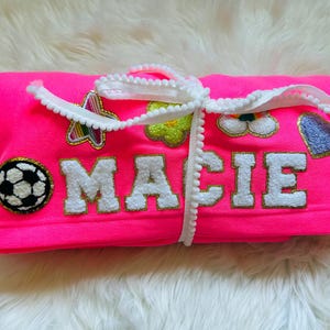 Könnte beinhalten: Eine rosa Fleece-Decke mit dem Namen "MACIE", der in Weiß mit goldener Umrandung gestickt ist. Die Decke hat verschiedene bunte Patches, darunter einen Fußball, einen Stern, eine Blume und ein Herz.