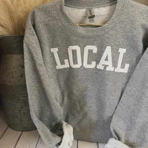 Puede incluir: Sudadera gris con la palabra "LOCAL" impresa en blanco en la parte delantera.