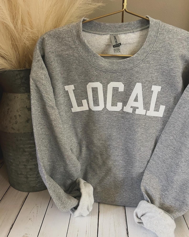 Puede incluir: Sudadera gris con la palabra "LOCAL" impresa en blanco en la parte delantera.