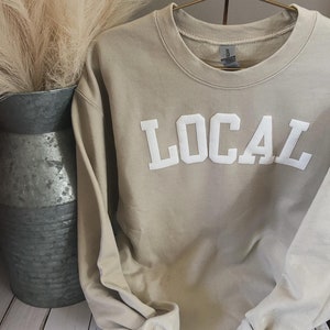 Puede incluir: Una sudadera beige con la palabra "LOCAL" impresa en blanco en la parte delantera. La sudadera est&aacute; colgada de una percha.