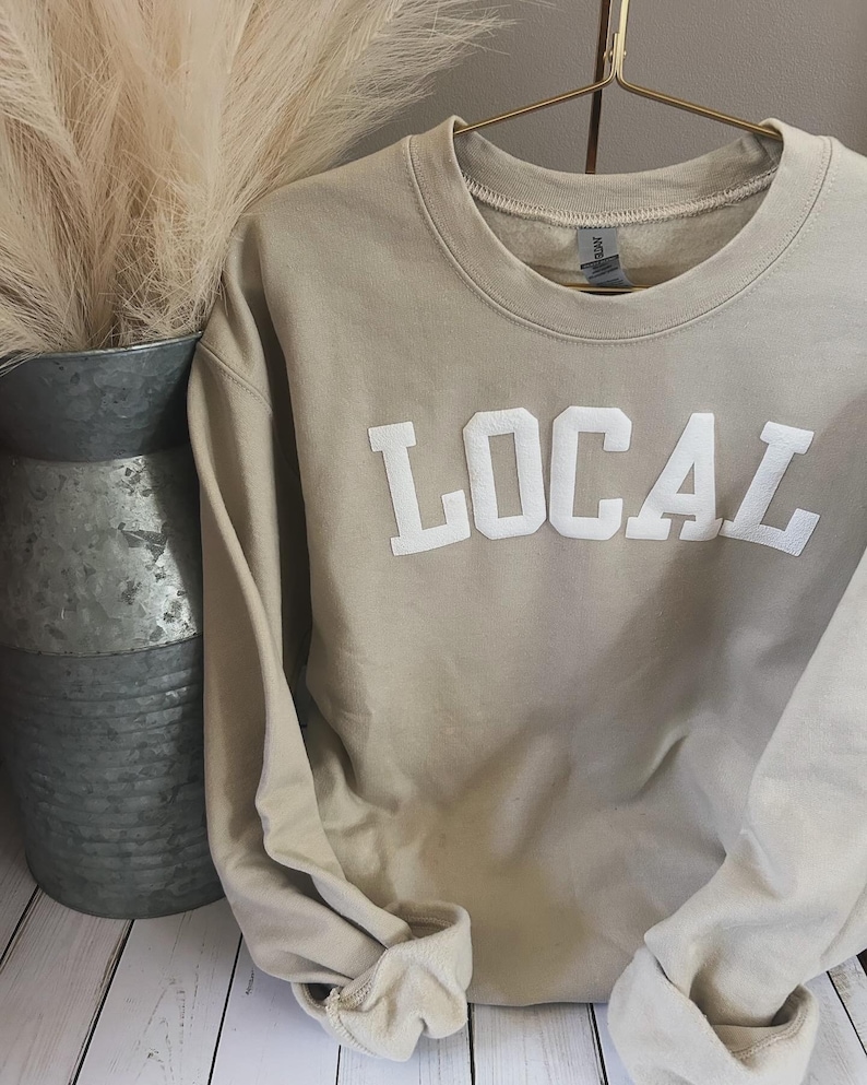Puede incluir: Una sudadera beige con la palabra "LOCAL" impresa en blanco en la parte delantera. La sudadera est&aacute; colgada de una percha.