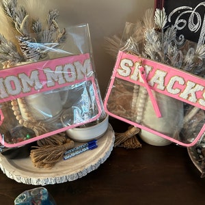 May include: Two clear plastic zippered pouches with pink fabric trim. One pouch has the text "MOM MOM" in white letters with gold glitter. The other pouch has the text "SNACKS" in white letters with gold glitter.