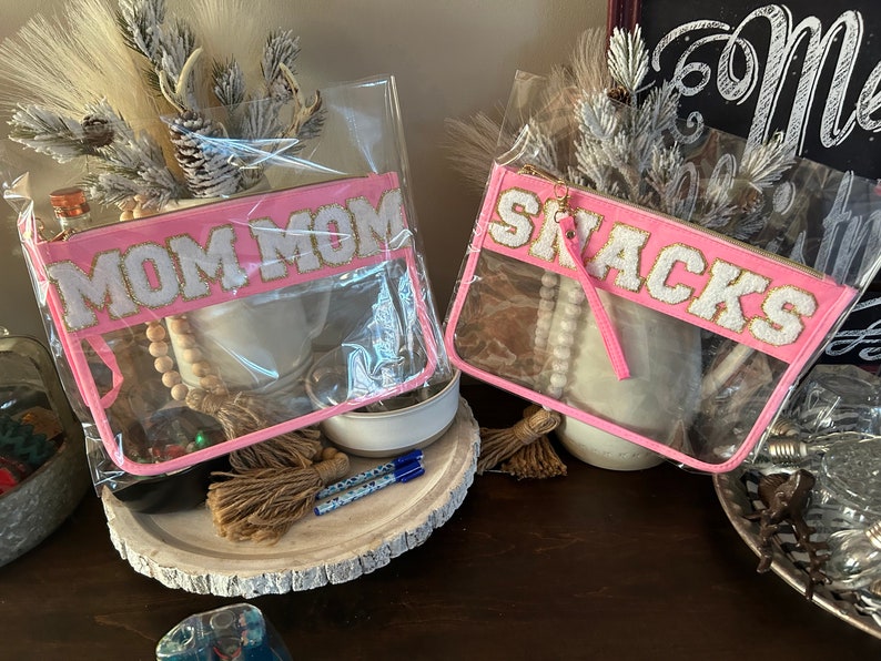 May include: Two clear plastic zippered pouches with pink fabric trim. One pouch has the text "MOM MOM" in white letters with gold glitter. The other pouch has the text "SNACKS" in white letters with gold glitter.