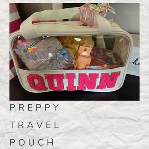 May include: A clear plastic travel pouch with a white base and a pink and gold "QUINN" name on the front. The pouch is filled with various items, including a pink and gold birthday cake lip gloss, a pink lip balm, and a gold hair clip.