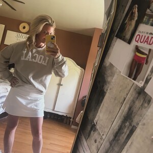 Puede incluir: Una mujer lleva una sudadera gris claro con la palabra "LOCAL" impresa en la parte delantera, una falda blanca y sandalias. Est&aacute; de pie frente a un espejo y sostiene un tel&eacute;fono amarillo.