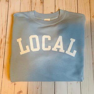 Puede incluir: Una sudadera azul claro con la palabra "LOCAL" impresa en blanco en la parte delantera.