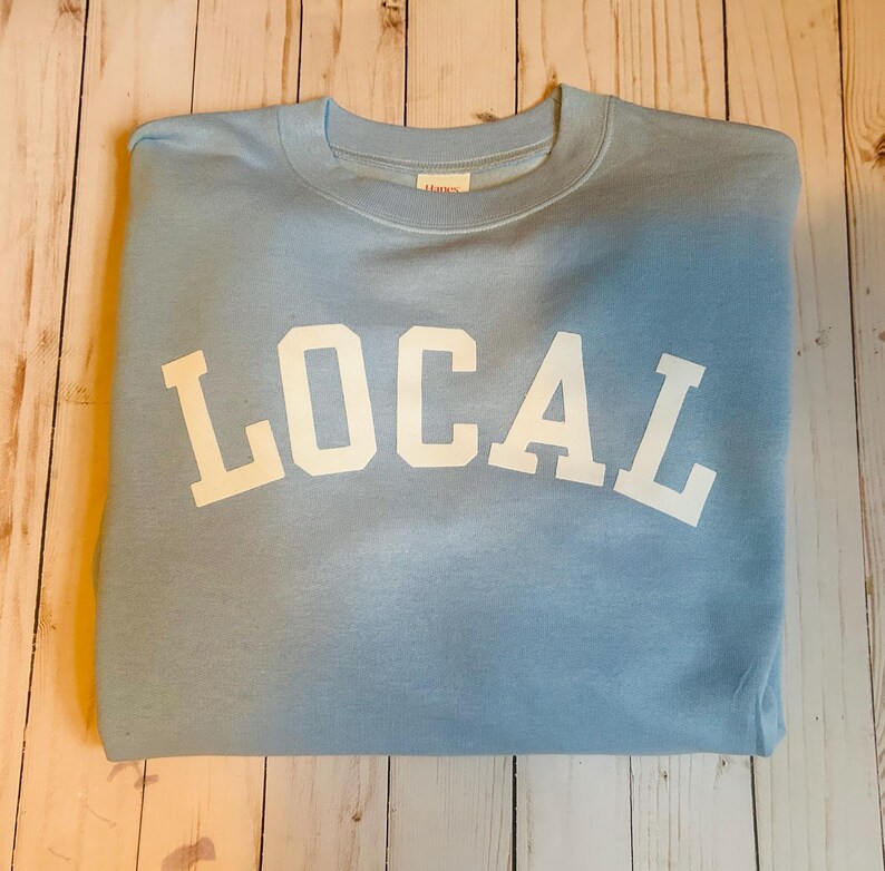 Puede incluir: Una sudadera azul claro con la palabra "LOCAL" impresa en blanco en la parte delantera.
