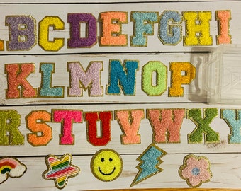 May include: A set of 26 colorful, glittery, felt letters with gold outlines, arranged in alphabetical order. The letters are in a variety of colors, including pink, blue, green, yellow, and purple. There are also five small, colorful, felt patches with various designs, including a rainbow, a star, a smiley face, a lightning bolt, and a flower.