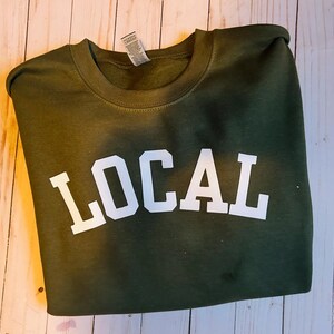 Puede incluir: Una sudadera verde con la palabra "LOCAL" impresa en blanco en la parte delantera.