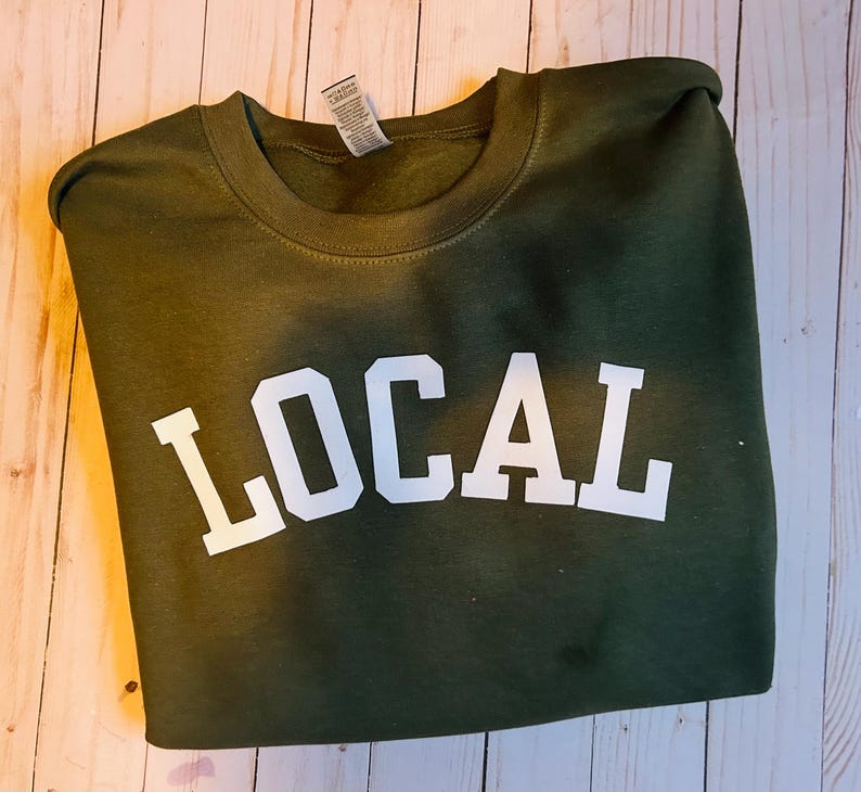 Puede incluir: Una sudadera verde con la palabra "LOCAL" impresa en blanco en la parte delantera.