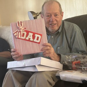 May include: A person holding a red and white striped gift box with a gold "DAD" label. The box is sitting on a white box.
