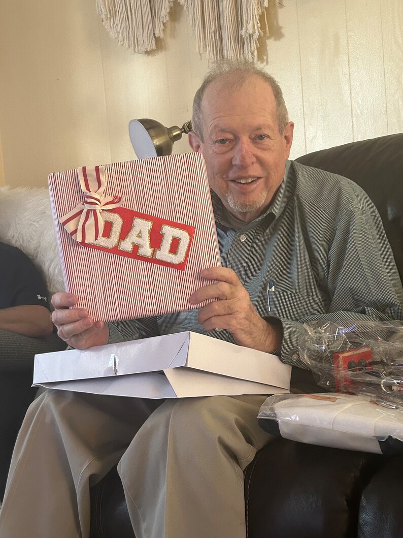 May include: A person holding a red and white striped gift box with a gold "DAD" label. The box is sitting on a white box.