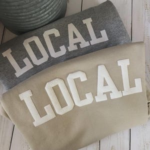 Puede incluir: Dos sudaderas con la palabra "LOCAL" impresa en blanco. La sudadera de arriba es gris y la de abajo es beige.