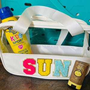 Può includere: Una borsa tote bianca, trasparente e con cerniera con un manico bianco. La parola "SUN" è scritta con lettere colorate e glitterate. All'interno della borsa ci sono due bottiglie di crema solare, una gialla e una marrone.