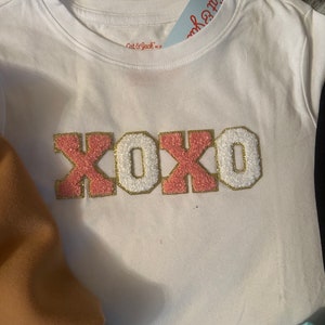 May include: White t-shirt with a pink and gold "XOXO" graphic. The shirt is made of a soft, comfortable material and has a crew neck.