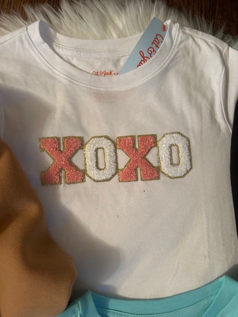 May include: White t-shirt with a pink and gold "XOXO" graphic. The shirt is made of a soft, comfortable material and has a crew neck.