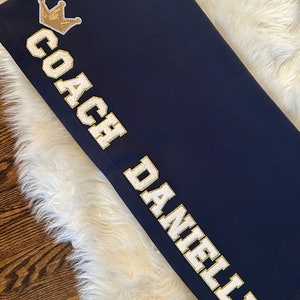 May include: Navy blue blanket with white text that reads "Coach Danielle" and a gold crown.