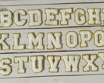 Può includere: Lettere in feltro bianco con contorni dorati, dalla A alla Z, disposte su tre righe. Le lettere sono maiuscole e hanno uno stile classico e universitario.
