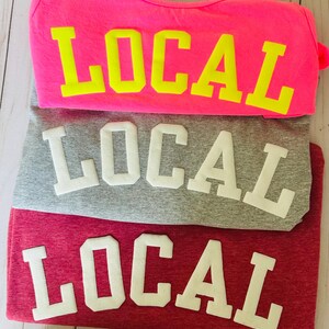 Puede incluir: Tres camisetas apiladas una encima de la otra. La camiseta de arriba es de color rosa intenso con la palabra "LOCAL" en letras may&uacute;sculas amarillas. La camiseta del medio es gris con la palabra "LOCAL" en letras may&uacute;sculas blancas. La camiseta de abajo es granate con la palabra "LOCAL" en letras may&uacute;sculas blancas.
