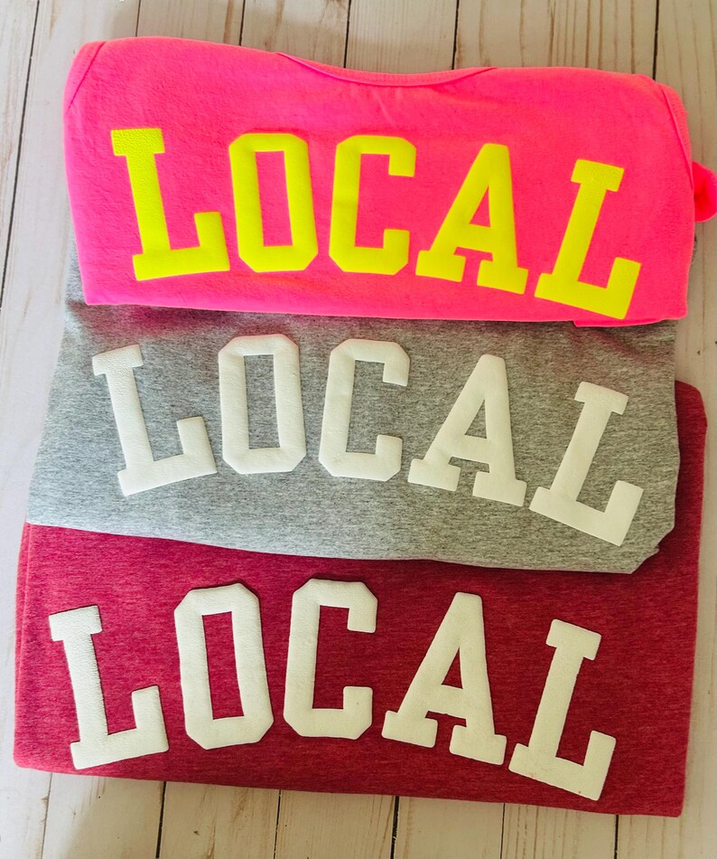 Puede incluir: Tres camisetas apiladas una encima de la otra. La camiseta de arriba es de color rosa intenso con la palabra "LOCAL" en letras may&uacute;sculas amarillas. La camiseta del medio es gris con la palabra "LOCAL" en letras may&uacute;sculas blancas. La camiseta de abajo es granate con la palabra "LOCAL" en letras may&uacute;sculas blancas.