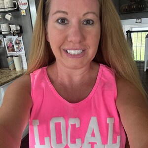 Puede incluir: Una mujer con una camiseta de tirantes rosa con la palabra "LOCAL" impresa en blanco en la parte delantera. Sonr&iacute;e y tiene el pelo largo y rubio.