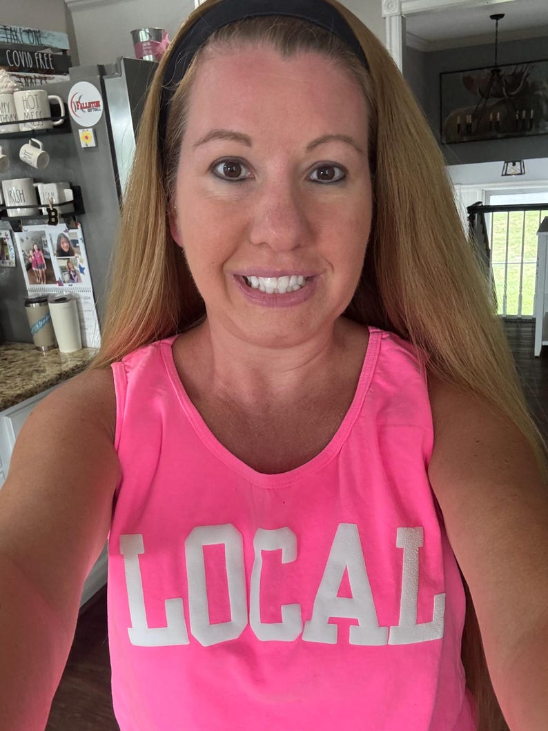 Puede incluir: Una mujer con una camiseta de tirantes rosa con la palabra "LOCAL" impresa en blanco en la parte delantera. Sonr&iacute;e y tiene el pelo largo y rubio.