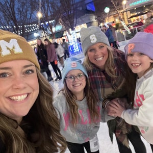 May include: Four people wearing winter hats with letters on them are smiling at the camera. The hats are gold, blue, light gray, and purple. They are standing on an ice rink.