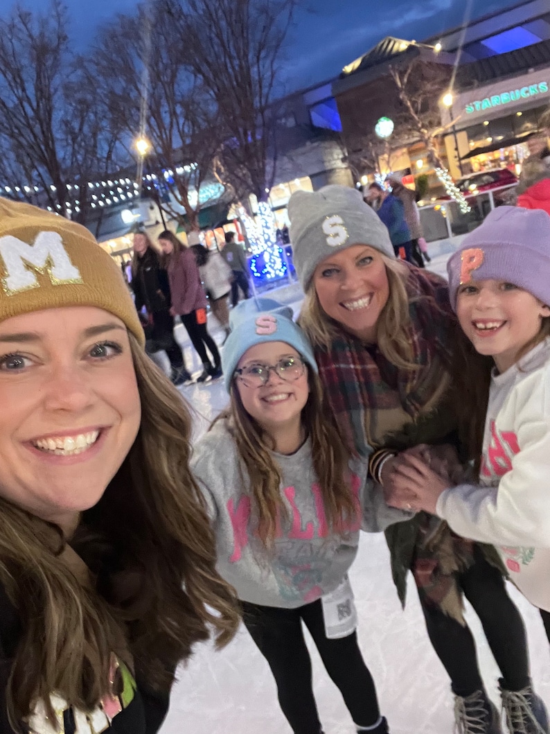 May include: Four people wearing winter hats with letters on them are smiling at the camera. The hats are gold, blue, light gray, and purple. They are standing on an ice rink.